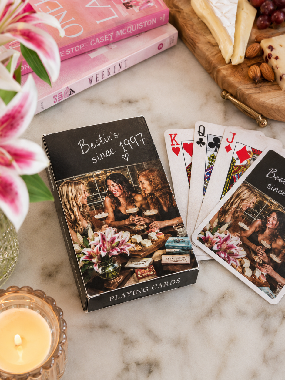 Playing cards with a photo of three women and text 'Besties since 1997' on a marble surface with books and flowers.